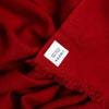 A close-up of a deep red cashmere scarf with fringed edges and a small white label sewn onto one corner.