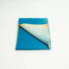 A folded blue cashmere item, likely a scarf or throw blanket, shown on a plain white background. One corner is turned over to reveal a cream-colored reverse side with yellow fringe.